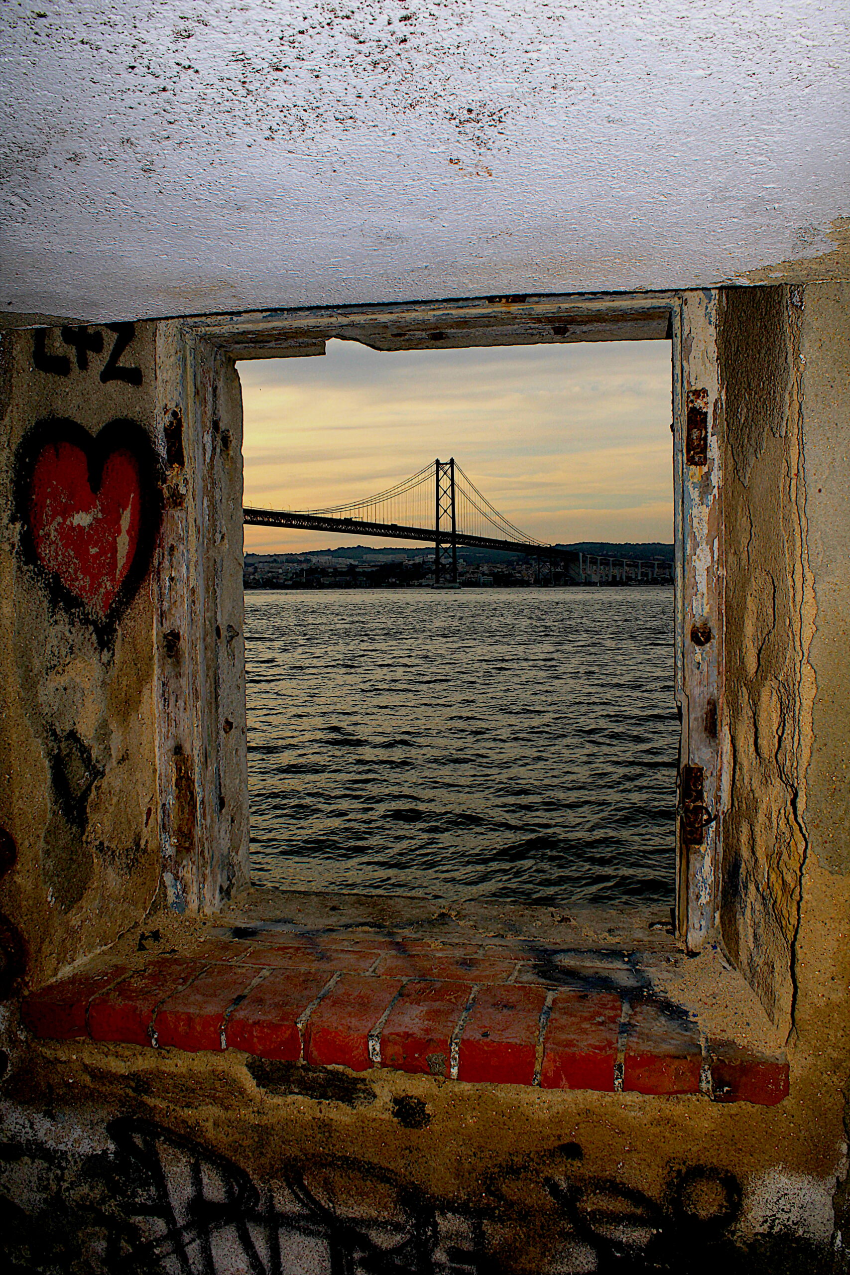 Perspetiva da Ponte 25 de Abril vista através de janela grafitada — contraste entre urbano e monumental